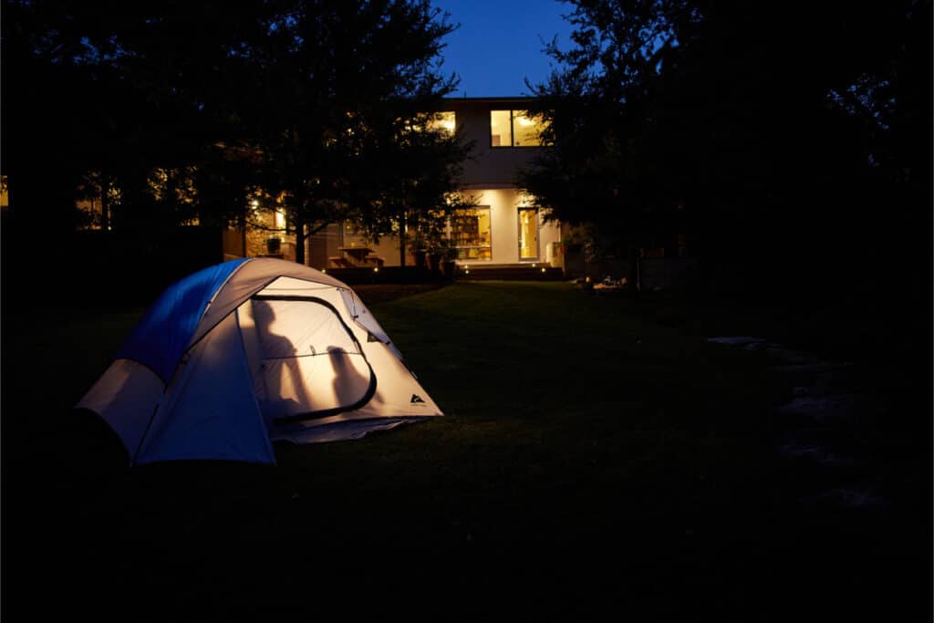 A Camping Tent Illuminated From Within Is Pitched On A Lawn At Twilight, With A Modern House Visible In The Background. The House's Interior Lights Cast A Warm Glow, Contrasting With The Darkening Sky And Surrounding Trees.