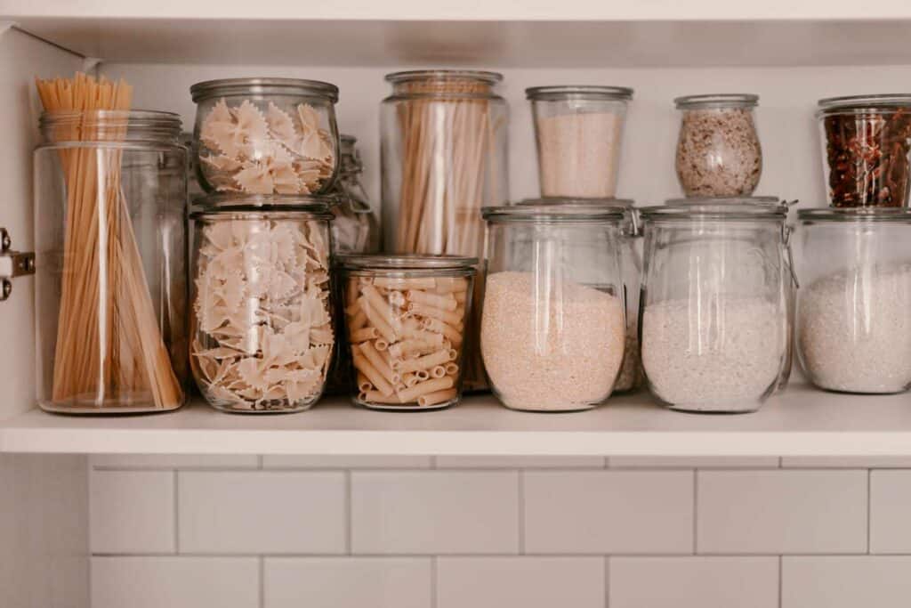 A Neatly Organized Kitchen Shelf Displaying Various Types Of Pasta And Grains Stored In Clear Glass Jars. The Jars Contain Spaghetti, Farfalle, Penne, Rice, And Other Dry Goods, All Arranged In A Clean And Orderly Manner Against A White Tiled Background.