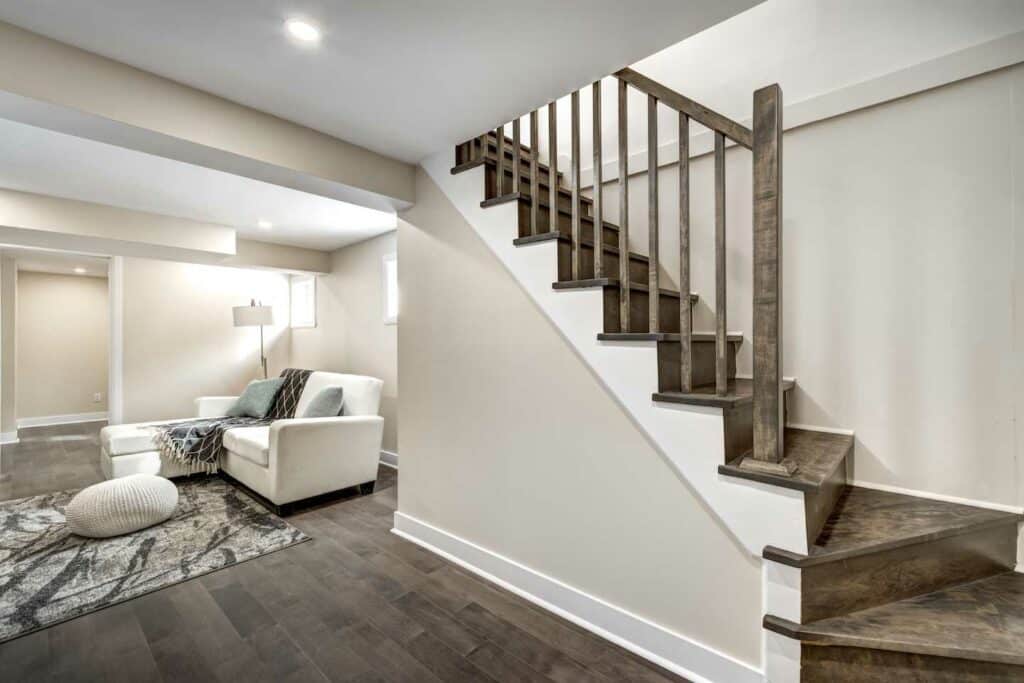 Modern Basement Interior With Light Beige Walls, Dark Wood Flooring, And A Cozy Seating Area Featuring A White Armchair And A Patterned Rug Next To A Stylish Staircase With Wooden Railings.