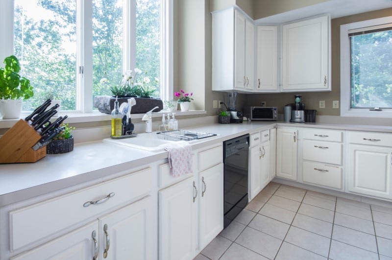 A Bright Kitchen With White Cabinets, A Large Window With Potted Plants, A Countertop With A Knife Block, A Dish Rack, And Various Kitchen Appliances.