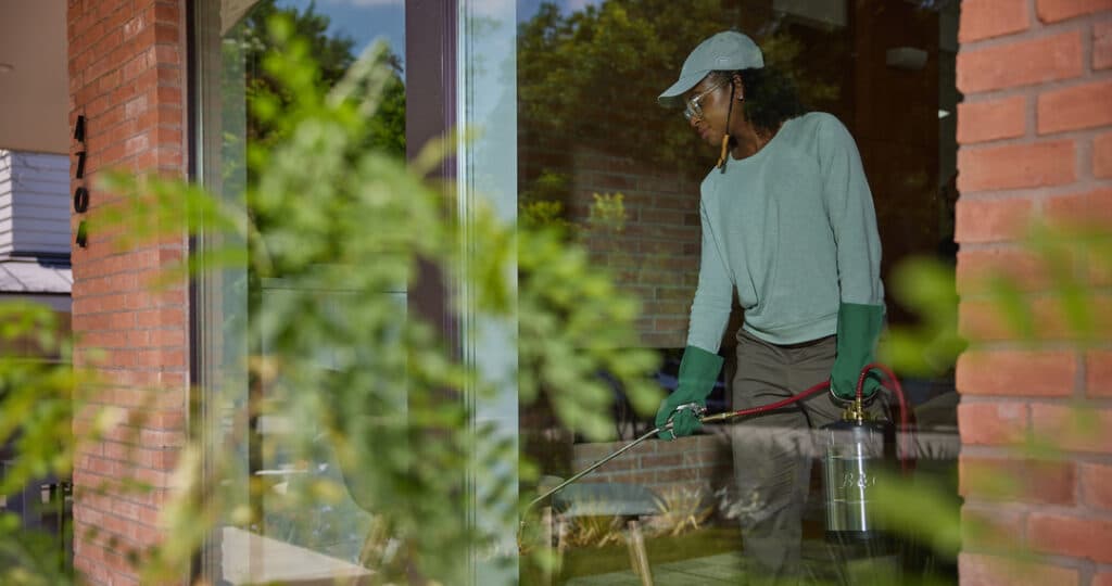 Aptive Pest Control Specialist Wearing Green Gloves And A Green Cap, Spraying Pesticide Near A Brick House With Address Number 4704 Visible.