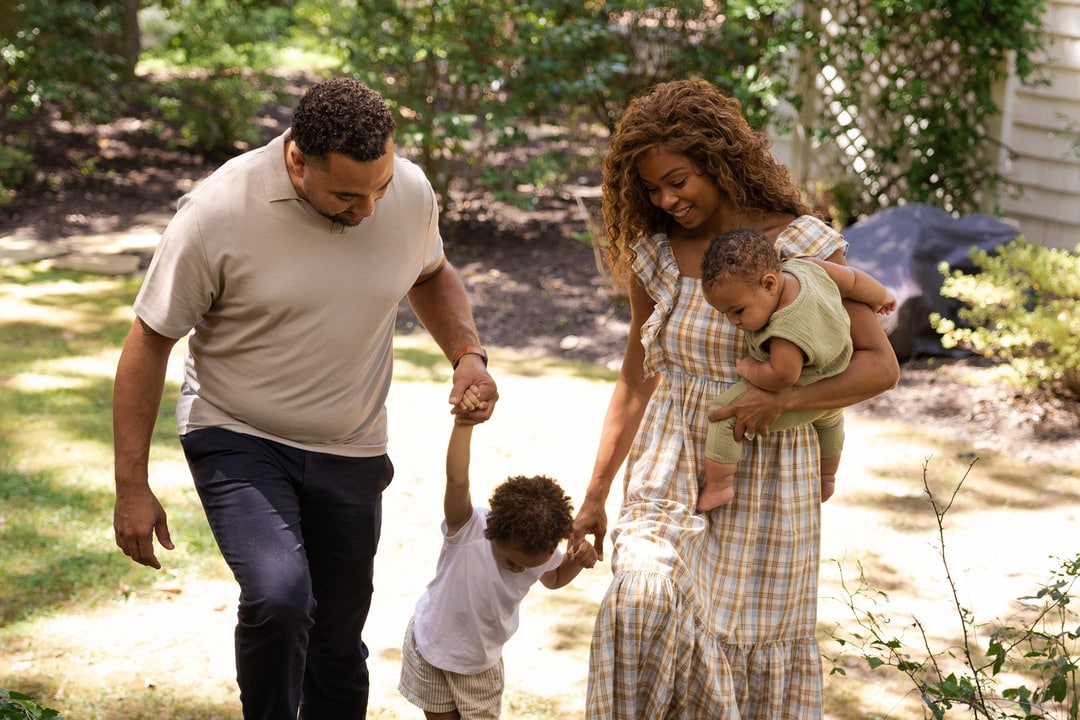 A Family Of Four, With The Parents Holding Hands With Their Young Children, Walks Through A Garden.