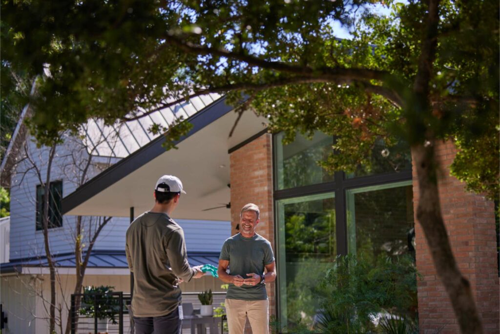 An Image Showing A Homeowner Smiling And Talking To An Aptive Pest Control Specialist In His Yard. The Specialist, Wearing A Company Cap And Shirt, Is Handing Over Some Documents To The Homeowner. They Stand In Front Of A Modern House With Large Windows And Lush Greenery, Under The Shade Of A Large Tree.