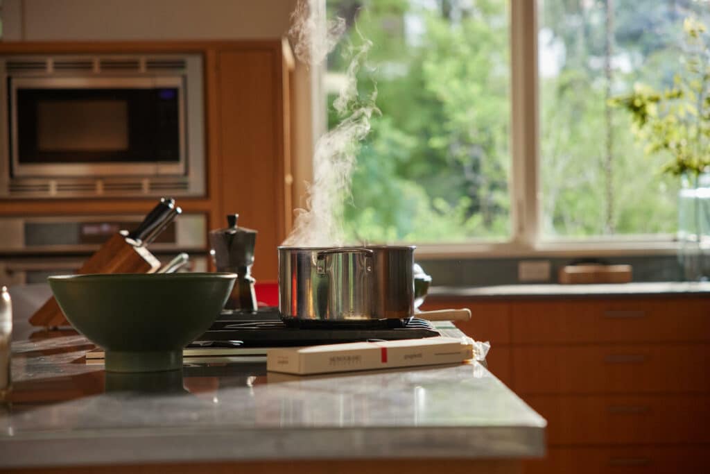 A Modern Kitchen With A Pot Of Steaming Water On The Stove, A Green Bowl, A Knife Block, And A Coffee Maker On The Countertop. The Background Features Large Windows With A View Of Greenery Outside, Letting In Natural Light.