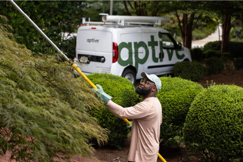 An Aptive Pest Control Specialist Using A Pole To Treat A Tree, With An Aptive Van Parked In The Background.