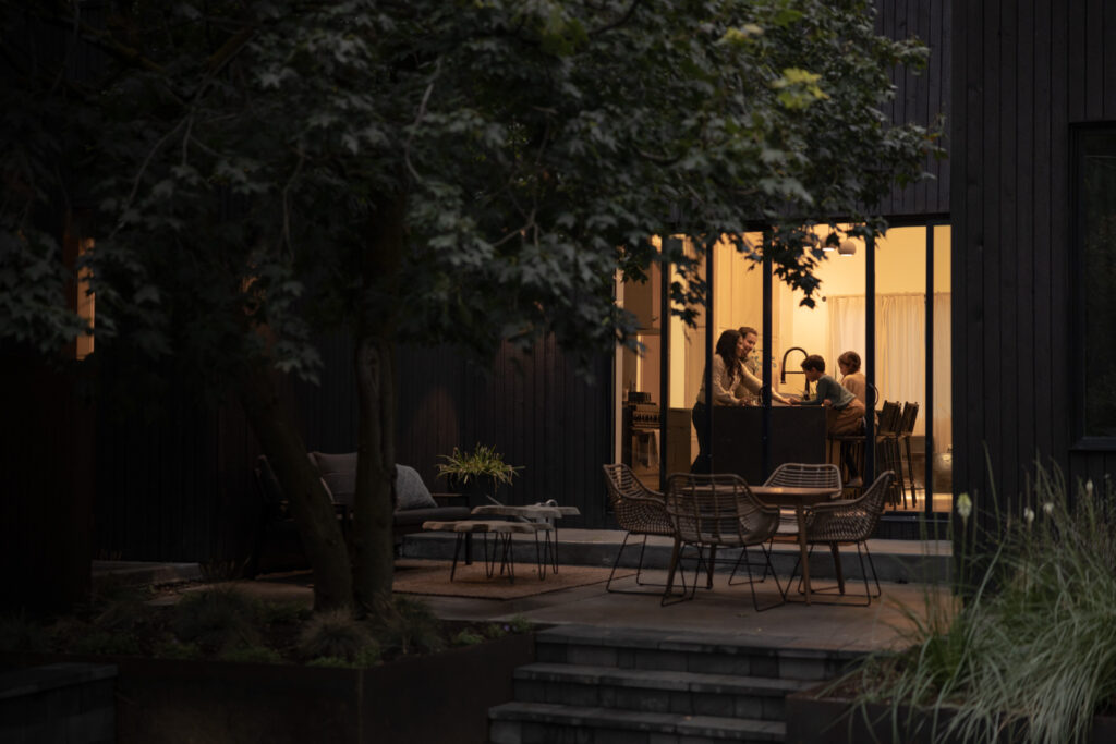 A Warm Evening View Of A Modern Home, Looking Through Large Windows Into A Kitchen Where A Family Of Four Is Gathered Around A Kitchen Island. Outside, The Patio Area Is Surrounded By Lush Greenery And Outdoor Furniture.