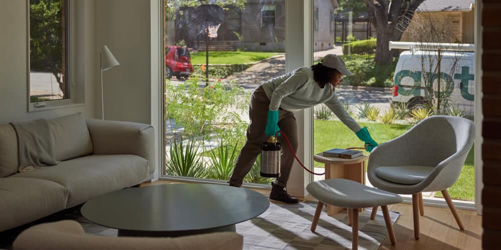 An Aptive Pest Control Specialist Wearing Gloves And A Cap, Spraying Inside A Modern Living Room With A View Of A Garden And An Aptive Van Outside.