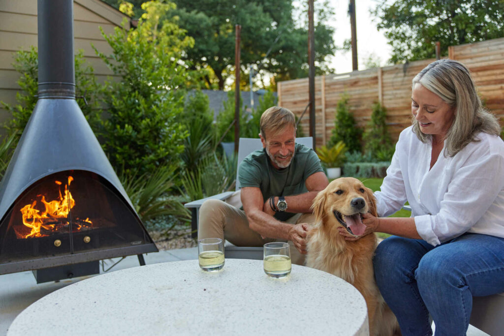 A Smiling Man And Woman Petting A Golden Retriever Next To A Round Outdoor Fireplace With Flames, Sitting In A Cozy Backyard With Lush Greenery.