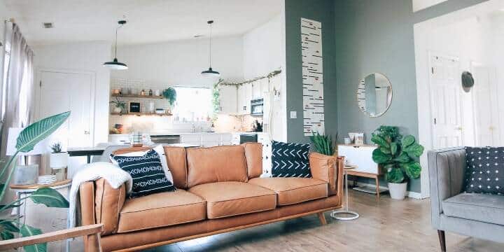 A Modern Living Room With A Tan Leather Sofa, Patterned Cushions, And Various Green Plants. The Room Opens Into A Bright, Stylish Kitchen With Hanging Lights And Wooden Countertops.