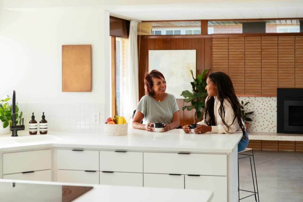 Two Women Sitting And Talking At A White Kitchen Island With A Bowl Of Fruit And Plants In The Background.