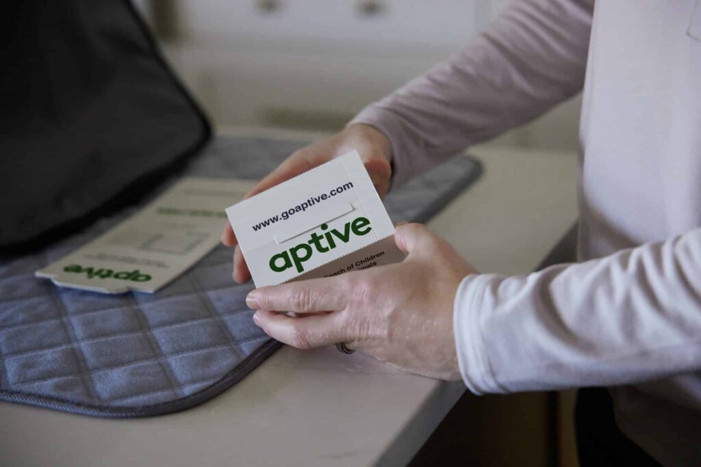 Close-Up Of Hands Holding A White Card With The Text Aptive, Over A Gray Padded Surface.