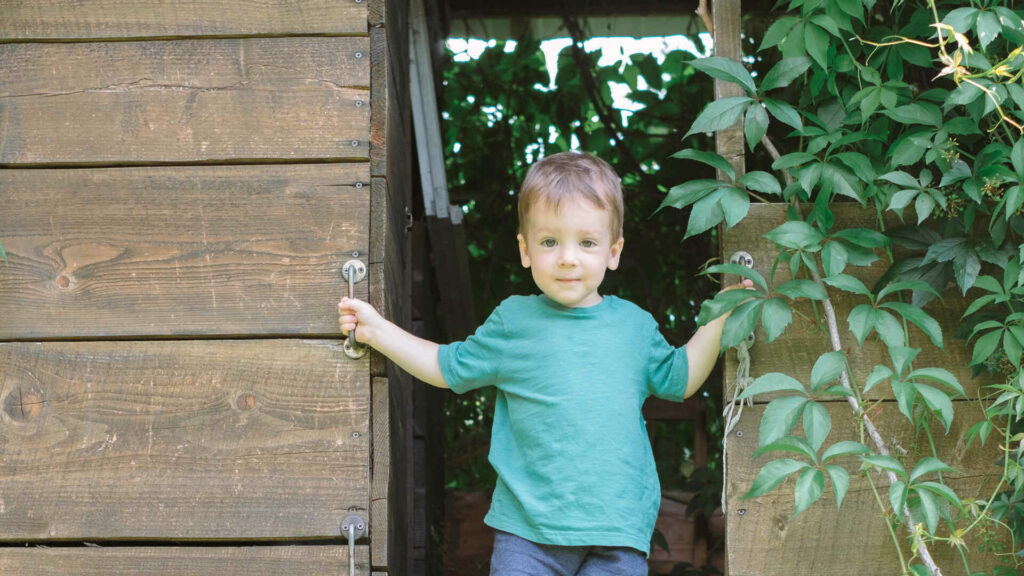 A Young Boy With Light Brown Hair Wearing A Green Shirt Standing In The Doorway Of A Wooden Structure, Surrounded By Green Foliage.