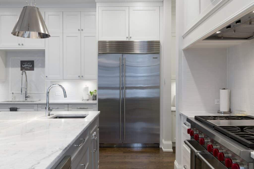 A Modern Kitchen Featuring White Cabinets, A Large Stainless Steel Refrigerator, A Marble Countertop Island With A Sink, And A Professional-Grade Stove With Red Knobs.