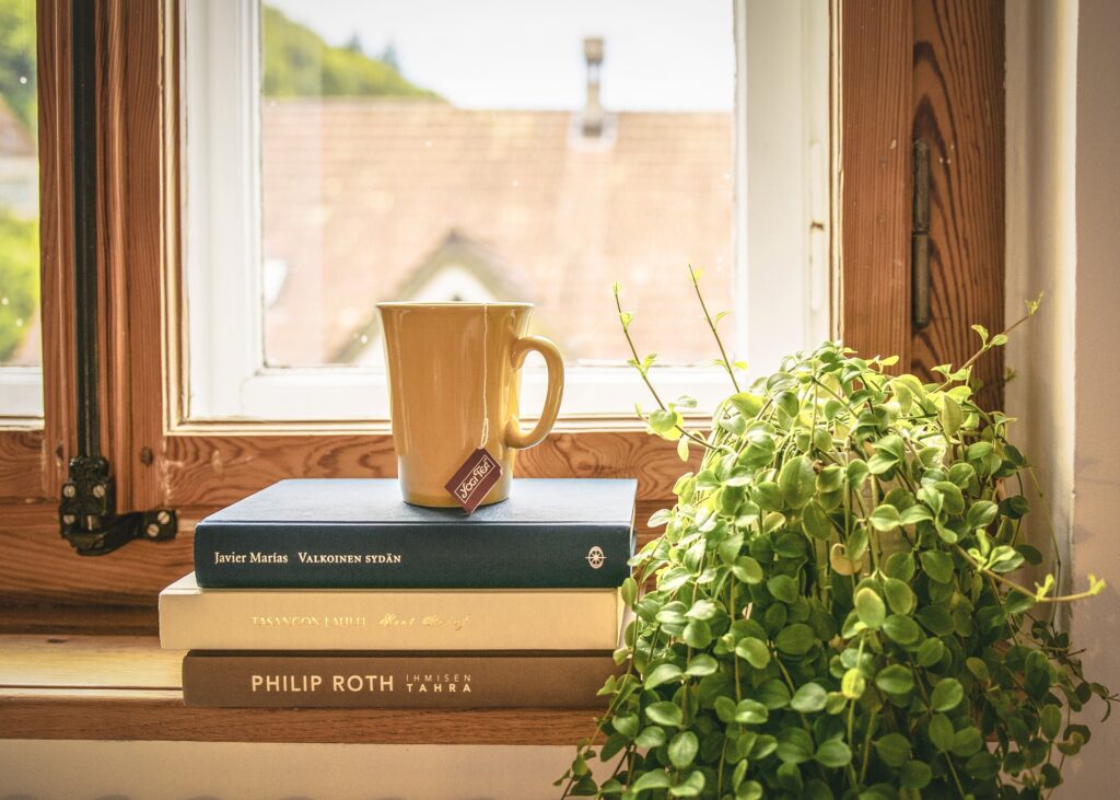 A Cozy Indoor Scene Featuring A Stack Of Three Books On A Wooden Windowsill. On Top Of The Books Rests A Yellow Ceramic Mug With A Tea Bag Label Hanging Over The Side. A Lush Green Houseplant Is Placed Beside The Stack, And The Window Behind Them Shows A Blurred View Of Rooftops And Greenery Outside.