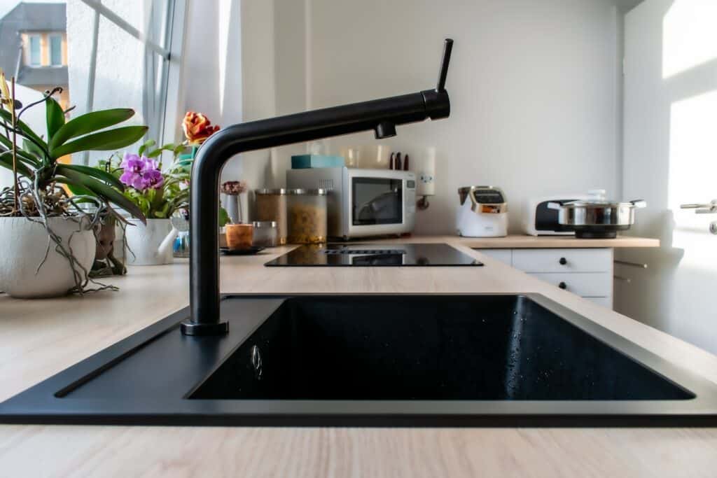 A Modern Kitchen Interior Showcasing A Sleek Black Sink With A Matte Black Faucet. The Kitchen Features A Wooden Countertop With Various Appliances Like A Microwave And Coffee Maker, And Is Decorated With Multiple Indoor Plants And A Small Fruit Bowl, Adding A Touch Of Nature And Color To The Space. Natural Light Streams In Through A Window, Illuminating The Area.