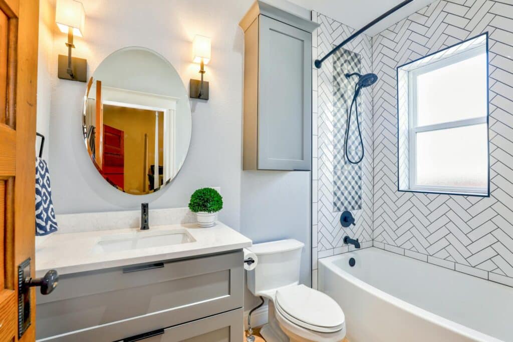 A Well-Lit Modern Bathroom Featuring A White Marble Countertop With A Rectangular Sink, A Large Circular Mirror Framed By Stylish Wall Lamps, And A Blue And White Patterned Shower Area With A Sliding Glass Door. The Room Has Subtle Accents Like A Small Potted Plant And A Blue Towel, Contributing To A Clean And Inviting Atmosphere.