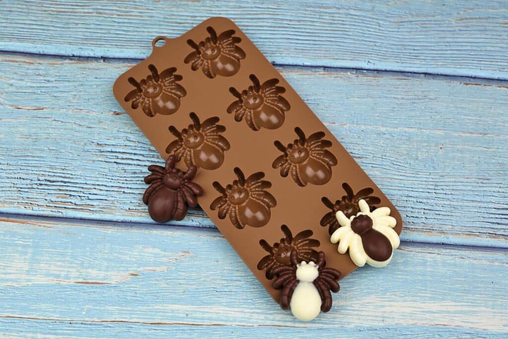 A Silicone Chocolate Mold Shaped Like Spiders, With Chocolate Spider Figures In Brown And White Placed On A Blue Wooden Surface.