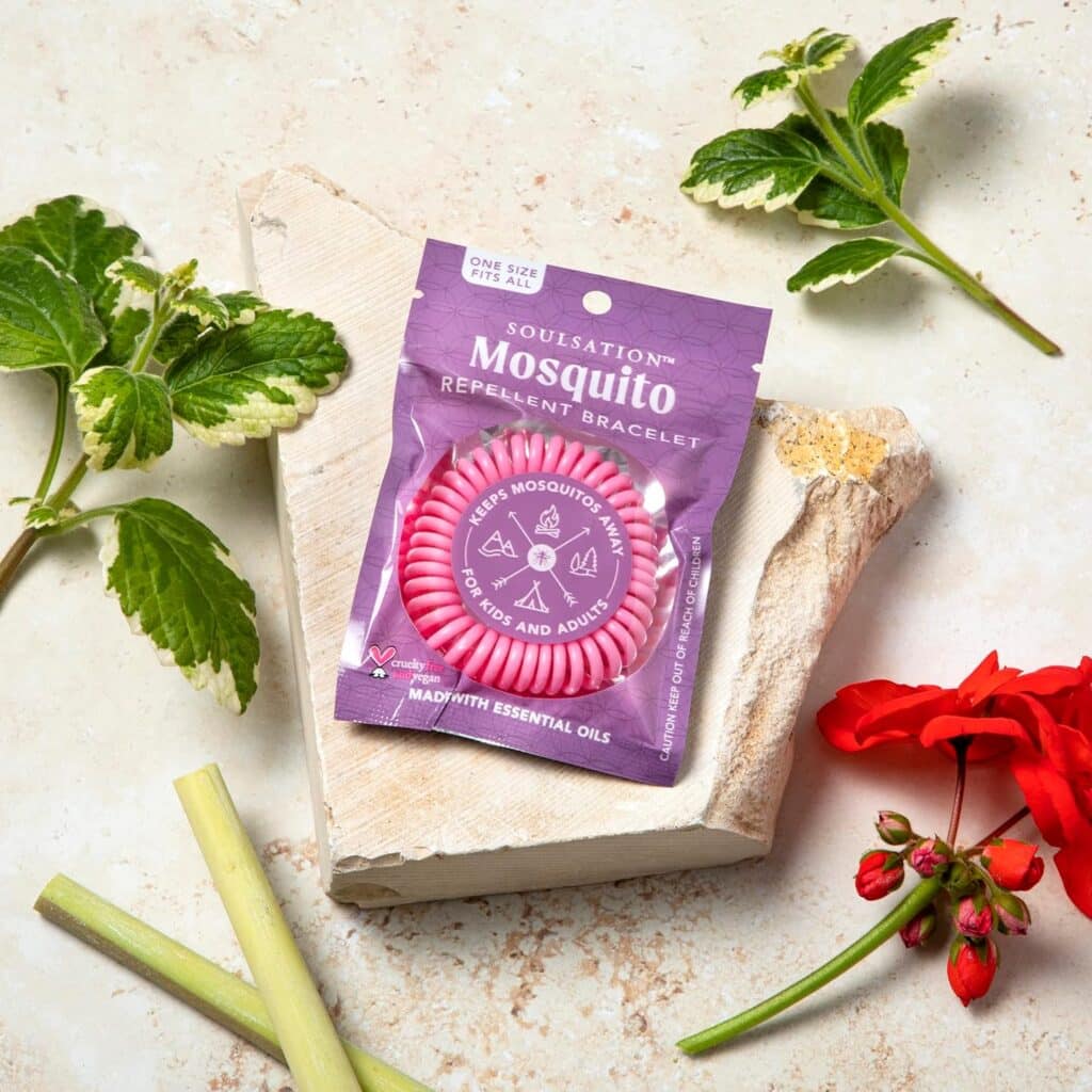 A Pink Soulsation Mosquito Repellent Bracelet In Its Packaging, Surrounded By Natural Elements Such As Geranium Flowers, Lemongrass, And Green Leaves.